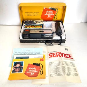 May include: A vintage Kodak Pocket Instamatic 20 camera kit. The kit includes the camera, flashcubes, a flash attachment, and instruction manuals. The camera is black and silver, with the Kodak logo. The box is yellow and black.
