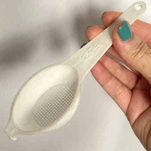 Puede incluir: Un colador de té Fuller de plástico blanco con un área de filtrado perforada. El colador tiene un mango largo con un agujero para colgar. El colador se sostiene en una mano con esmalte de uñas turquesa.