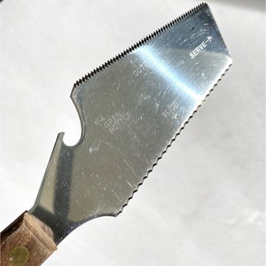 Op de afbeelding: Een zilveren multifunctioneel keukenhulpmiddel met een houten handvat. Het gereedschap heeft een gekartelde rand om te snijden, een flesopener en markeringen met de tekst "OPEN BOTTLE", "CUT", "SLICE" en "SERVE".