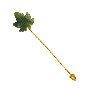 Puede incluir: Un broche verde tallado en forma de hoja, con un tallo dorado y un extremo decorativo. La hoja tiene vetas detalladas y una superficie texturizada. El broche está sobre un fondo blanco.