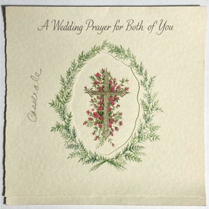 May include: Cream-colored wedding card with the text "A Wedding Prayer for Both of You." The card features a gold cross adorned with pink roses, surrounded by a green floral wreath. The name "Cassiole" is handwritten on the left side.