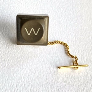 Può includere: Una spilla da bavero in plastica marrone con la lettera 'W' e una catena e chiusura dorate.