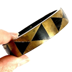Può includere: Un braccialetto rigido dorato e nero con un motivo geometrico. Il braccialetto è in metallo e ha una finitura lucida.