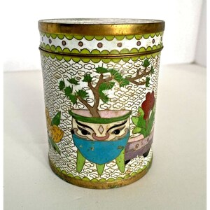 Puede incluir: Un recipiente cilíndrico de cloisonné con borde y base dorados. El cuerpo blanco presenta un borde verde y dorado, con un diseño de bonsái en una maceta azul y otros elementos florales. El recipiente mide aproximadamente 10 cm de alto.
