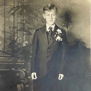 Puede incluir: Un niño pequeño con un traje negro, camisa blanca y corbata oscura. Lleva una flor de solapa con flores blancas. El niño está de pie frente a un fondo con un diseño de hojas.