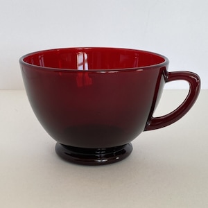 May include: A deep red glass teacup with a rounded bowl and a single handle. The cup has a small foot at the base.