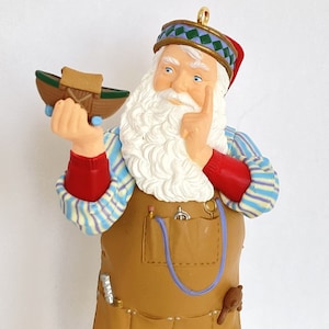 May include: A Santa Claus ornament with a white beard and a blue and green striped shirt. He is holding a small wooden boat in his hand and is wearing a brown leather apron with tools.