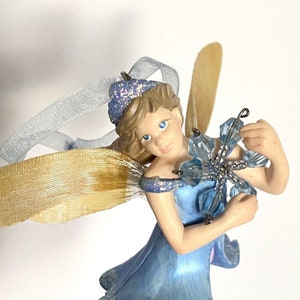 May include: A blue fairy figurine with a gold and white ribbon, holding a blue snowflake ornament. The fairy has blonde hair and blue eyes and is wearing a blue dress.