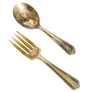 May include: An antique silver spoon and fork set against a white background. The spoon has a rounded bowl, and the fork has four tines. Both utensils show signs of age and wear, with tarnishing and discoloration.
