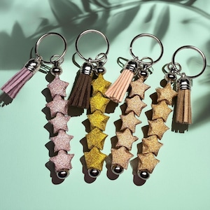 May include: Four keychains with star-shaped charms and tassel accents. The keychains feature silver rings, glittery star charms in pink, gold, and brown, and matching tassels. The background is a light teal color.