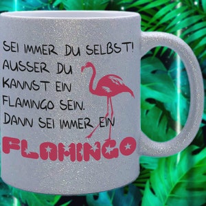 Puede incluir: Una taza de cerámica blanca con un acabado brillante presenta un flamenco rosa y el texto "SEI IMMER DU SELBST! AUSSER DU KANNST EIN FLAMINGO SEIN. DANN SEI IMMER EIN FLAHINGO"