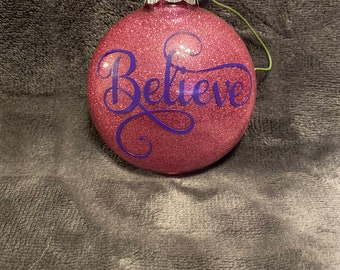 May include: Pink glitter Christmas ornament with a purple cursive "Believe" design.