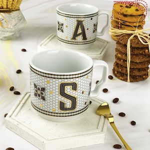 May include: Two white ceramic coffee mugs with black and gold mosaic designs. The mugs feature the letters "A" and "S" in gold. The mugs are on white hexagonal coasters. Coffee beans are scattered around the mugs.