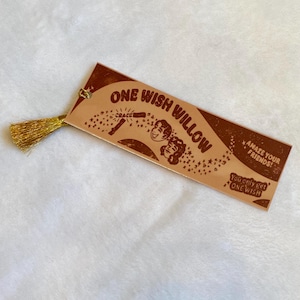 May include: A rectangular bookmark with a metallic gold finish and a gold tassel. The design features brown text reading ONE WISH WILLOW, AMAZE YOUR FRIENDS!, and You only get ONE WISH, along with illustrations of a wand, stars, and two faces.