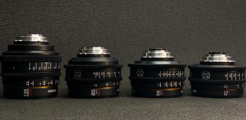 May include: Four black camera lenses with silver accents and markings. The lenses are arranged in a row from left to right, with the largest lens on the left and the smallest lens on the right. The lenses have markings indicating their focal lengths: 135mm, 85mm, 58mm, and 32mm.