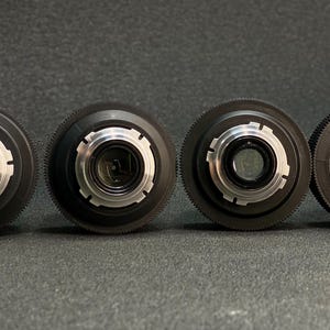 May include: Four black camera lenses with silver accents and adjustable aperture rings. The lenses are arranged in a row on a grey surface.