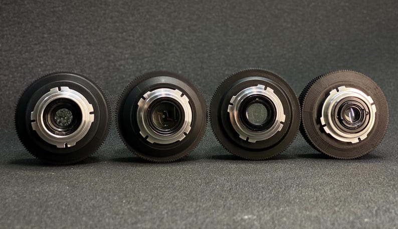 May include: Four black camera lenses with silver accents and adjustable aperture rings. The lenses are arranged in a row on a grey surface.