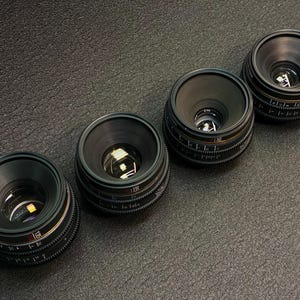 May include: Four black camera lenses with silver accents and markings. The lenses are arranged in a row from left to right, with the largest lens on the left and the smallest lens on the right.