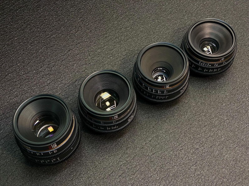 May include: Four black camera lenses with silver accents and markings. The lenses are arranged in a row from left to right, with the largest lens on the left and the smallest lens on the right.