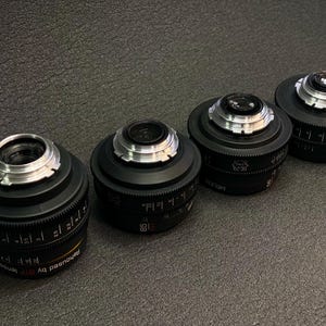 May include: Four black camera lenses with silver accents and markings. The lenses are arranged in a row from left to right, with the largest lens on the left and the smallest lens on the right.