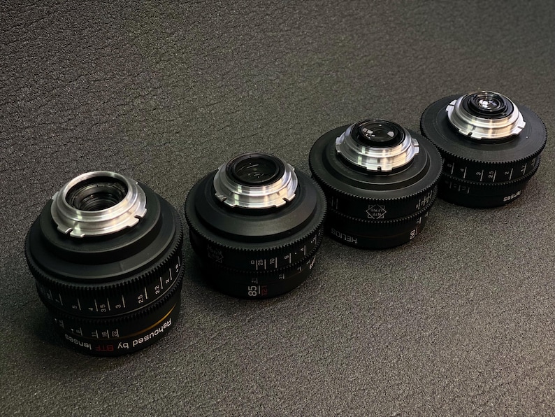 May include: Four black camera lenses with silver accents and markings. The lenses are arranged in a row from left to right, with the largest lens on the left and the smallest lens on the right.