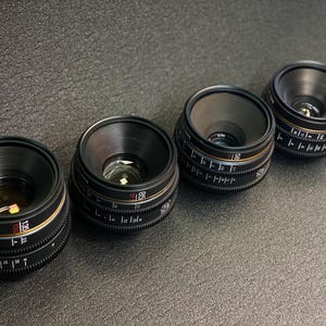 May include: Four black camera lenses with gold and red markings. The lenses are arranged in a row from left to right.