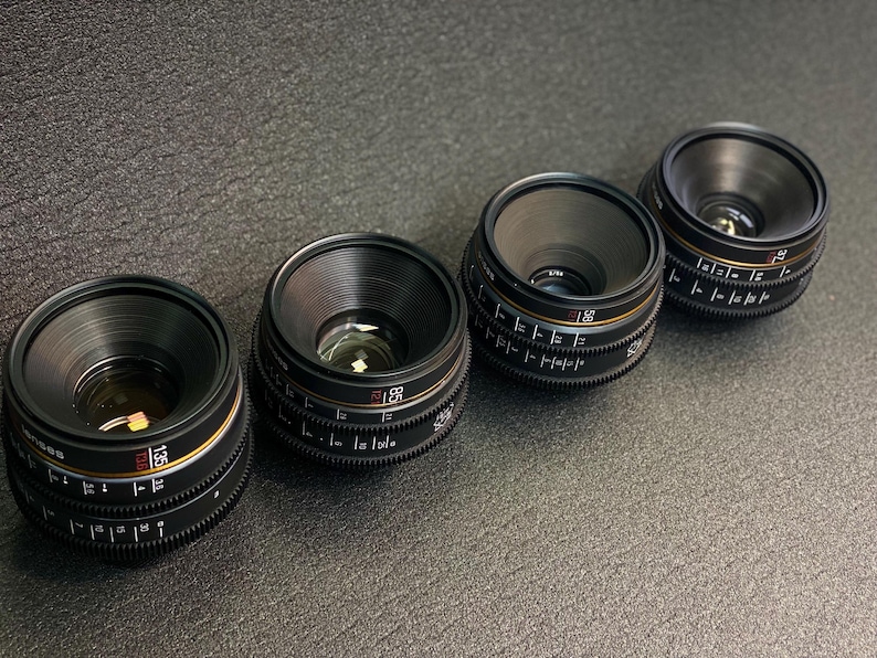 May include: Four black camera lenses with gold and red markings. The lenses are arranged in a row from left to right.