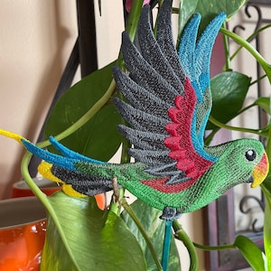 May include: A green, red, blue, and yellow embroidered parrot with black wings is attached to a green stick. The parrot is in flight and is perched on a green plant.