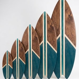 Puede incluir: Cinco tablas de servir de madera con forma de tabla de surf en tamaños 26x10cm, 40x15cm, 60x25cm, 80x30cm y 100x40cm. Cada tabla presenta una veta de madera marrón oscura, una incrustación central de resina azul verdosa y una base teñida.