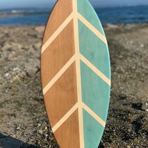 Könnte beinhalten: Ein Holz-Balanceboard mit einem geometrischen Design in Teal und Braun. Das Board ist surfboardförmig und steht auf einem Holzsockel.