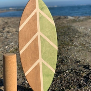 Könnte beinhalten: Ein hölzernes Balanceboard in Form eines Blattes mit einem geometrischen Muster in Grün und Braun. Das Board steht auf einem Holzsockel und befindet sich an einem Strand mit Kieselsteinen.
