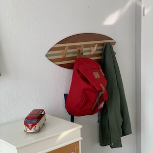 May include: A wooden surfboard-shaped coat rack with three hooks. A red backpack and a green jacket are hanging on the hooks. A red and white toy bus is on a white shelf.