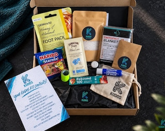 May include: A cardboard gift box containing a variety of items including a DermaV10 foot pack, a bag of Haribo Starmix, a Hawaiian Tropic sunscreen sachet, a packet of Kudos bath salts, an emergency blanket, a lip balm, and a cotton drawstring bag.
