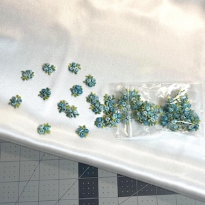 Könnte beinhalten: Eine Sammlung kleiner, hellblauer Stoffrosen-Applikationen mit grünen Blättern. Mehrere einzelne Blumenapplikationen sind verstreut, während sich ein größeres Bündel dieser floralen Elemente in einem durchsichtigen Plastikbeutel befindet.
