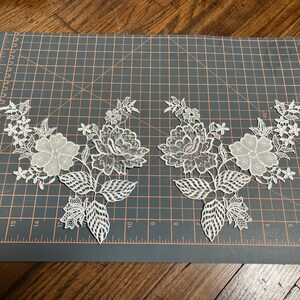 May include: Two white lace floral appliques. Each applique features a large flower, smaller blossoms, and detailed leaves. The appliques are arranged on a gray cutting mat with grid lines, ideal for sewing or crafting projects.