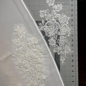 May include: Two white floral lace appliques featuring intricate rose and leaf embroidery on sheer mesh backing. One applique is placed on a white fabric surface, while the other rests on a gray cutting mat with inch markings.