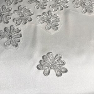 Könnte beinhalten: Ein weißer Stoff mit einer Reihe von gestickten silbernen Blumenmotiven. Die Blumenmuster sind in einer verstreuten Anordnung auf der glatten, einfarbig weißen Textiloberfläche verteilt und bilden ein wiederkehrendes Design.