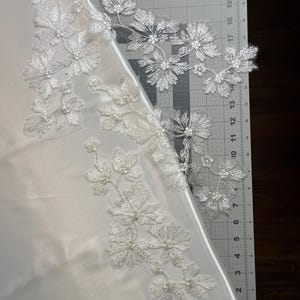 Könnte beinhalten: Weiße florale Spitzenapplikation mit aufwendiger Stickerei und kleinen Perlenakzenten, die auf einem glatten weißen Satinstoff liegt. Die Spitzenteile sind auf einer Schneidematte mit sichtbaren Markierungen von 2 bis 56 Zentimetern angeordnet.