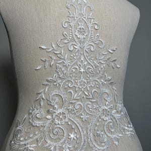 Könnte beinhalten: Weiße Spitzenapplikation mit floralem Muster und Pailletten. Die Applikation ist perfekt, um jedem Kleidungsstück einen Hauch von Eleganz zu verleihen.