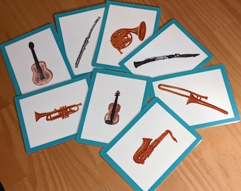 May include: A collection of flashcards featuring illustrations of musical instruments. The cards have a white background with a teal border. Instruments include a guitar, flute, French horn, clarinet, trumpet, violin, trombone, and saxophone.