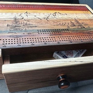 May include: A wooden cribbage board with a mountain scene inlay. The board has a drawer for storing cards and other game pieces. The drawer has a wooden knob.