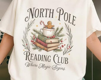 May include: Cream-colored t-shirt with the text "North Pole Reading Club Where Magic Begins." The graphic features a gingerbread man in a mug atop books, surrounded by greenery and berries. The shirt has short sleeves.