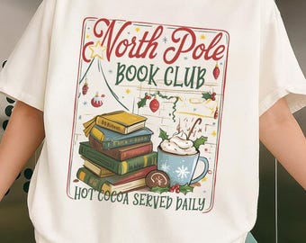 May include: Cream-colored t-shirt featuring a graphic for the "North Pole Book Club." The design includes a stack of books, a mug of hot cocoa, and festive holiday decorations. The text "Hot Cocoa Served Daily" is also included.