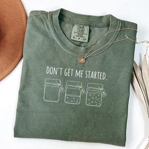 May include: An olive green t-shirt with the phrase "DON'T GET ME STARTED" printed in white. Below the text are three jar illustrations. A gold necklace with a pendant rests on the shirt. The shirt is folded on a white surface.