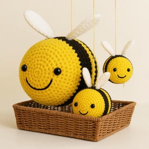 May include: A set of three crocheted bee ornaments. The bees are yellow with black stripes and white wings. They have smiling faces and are displayed in a brown wicker basket.