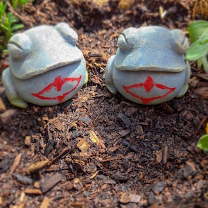 Puede incluir: Dos figuritas de rana grises con sonrisas pintadas de rojo, sentadas en un lecho de mantillo marrón.