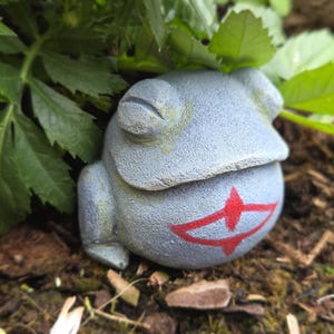 May include: A grey frog ornament with a red design, set amongst green leaves and brown mulch. The frog has a textured surface. The garden ornament is a charming addition to any outdoor space.