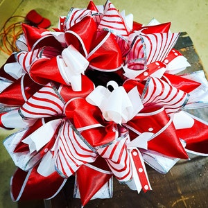 May include: A large, decorative bow featuring layers of red and white satin ribbons. Some ribbons have a striped pattern, while others are solid red or white. Silver trim accents the edges, creating a festive and eye-catching design.