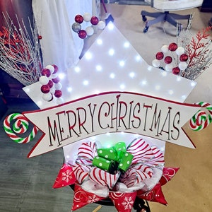 May include: A white star-shaped Christmas decoration with the words "MERRY CHRISTMAS" in red lettering. The decoration is adorned with red and white striped bows, candy canes, and decorative branches. The star is lit with small white lights.