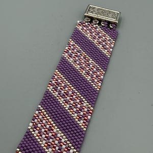 May include: A beaded bracelet featuring a purple base with diagonal stripes of white, rose gold, and gold beads. The bracelet has a silver-colored clasp. This handcrafted accessory is suitable for everyday wear.